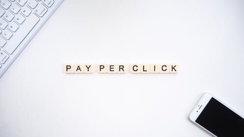 Pay per Click written in scrabble letters-white android phone-corner of keyboard-white surface